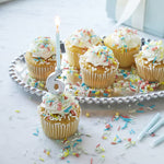 Cupcakes with white frosting and colorful sprinkles on a silver platter with a number 6 candle.