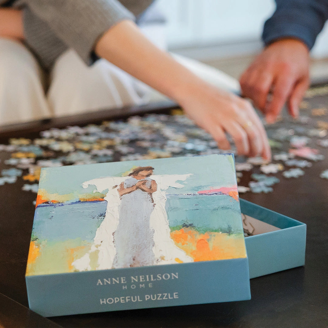 Jigsaw puzzle with a box featuring 'Anne Neilson Home' and 'Hopeful Puzzle' on a table with hands working on the puzzle.