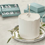 White candle with a lit candle on top, placed on a silver tray with green leaves, against a light background.