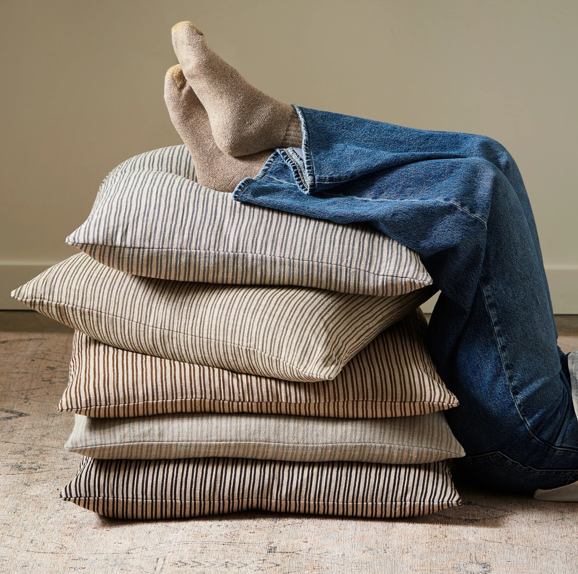 Person wearing blue jeans and brown socks sitting on a stack of striped pillows.
