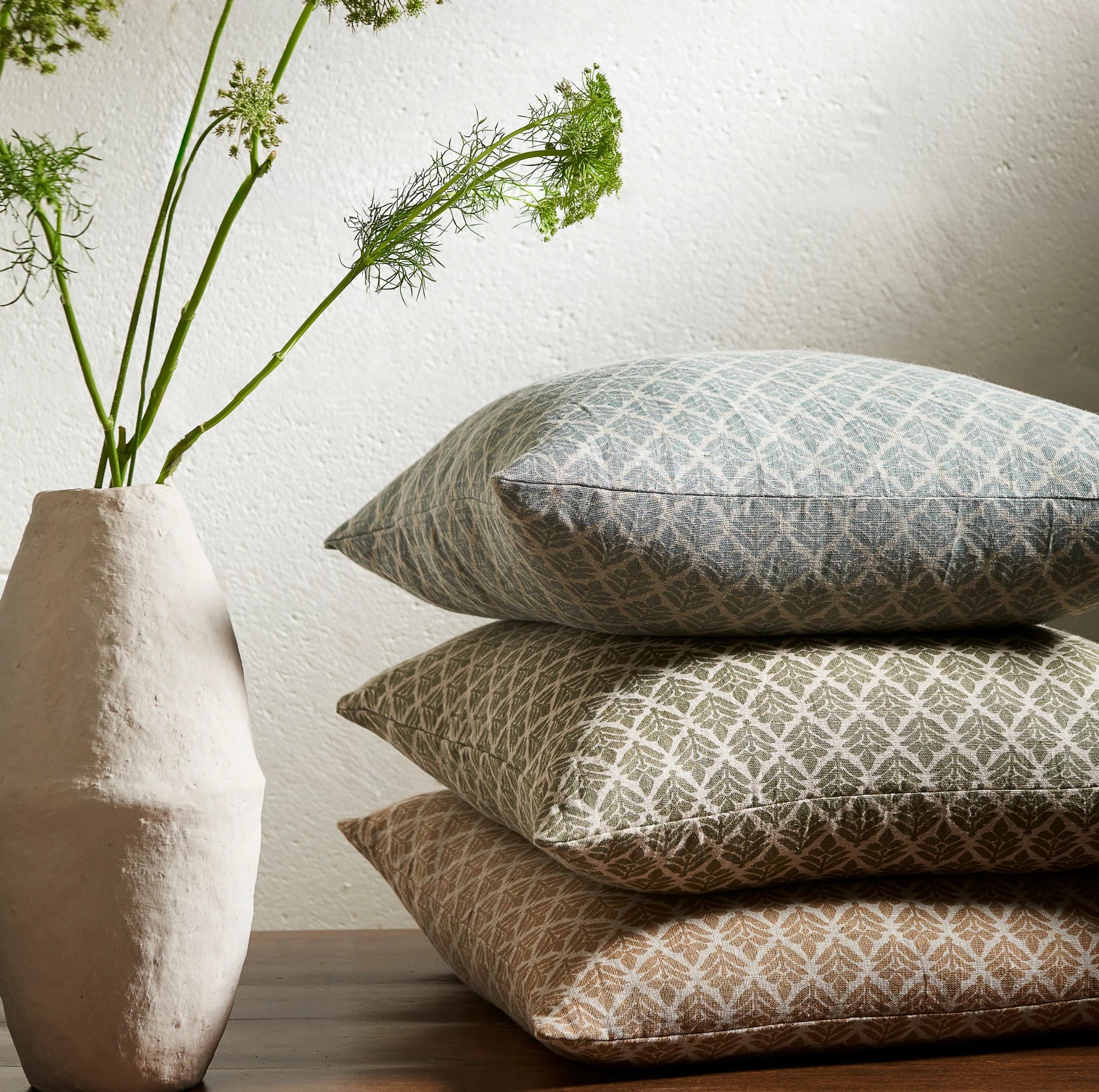 Stack of patterned pillows with a vase containing greenery on a neutral background