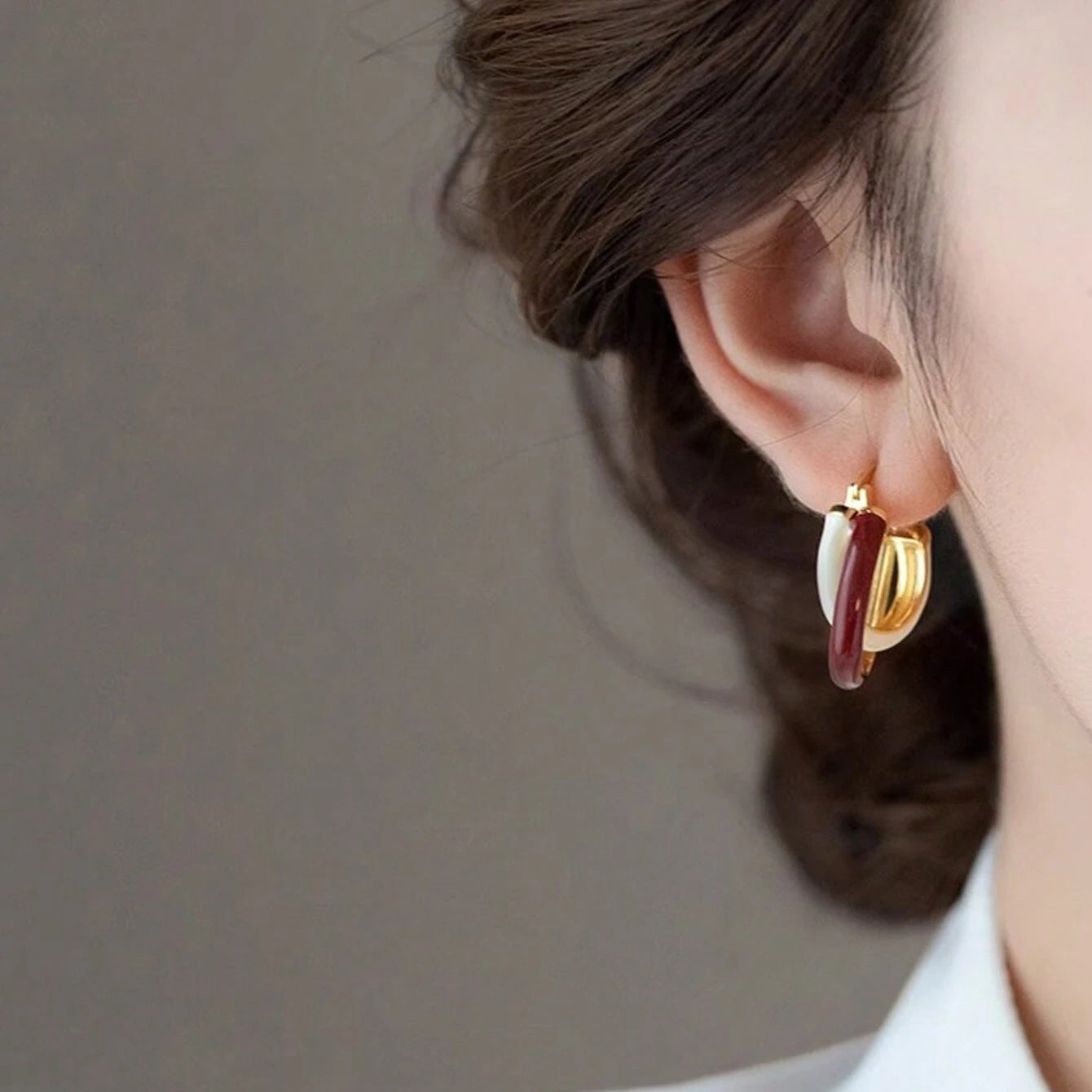 Close-up of a person wearing gold and burgundy hoop earrings against a neutral background.