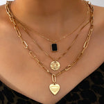 Model wearing a three-layer gold necklace with black pendant, coin charm, and heart charm layered neatly together, styled with a leopard-print top.