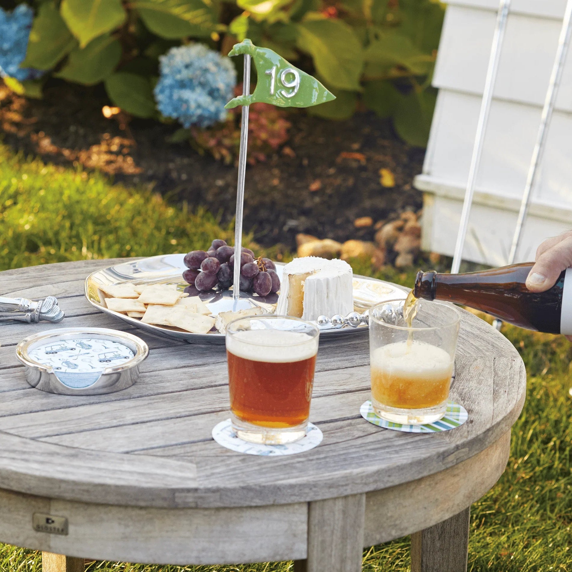 Outdoor setting with a wooden table, drinks, snacks, and a person pouring beer.