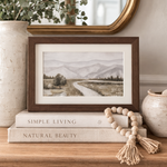 Framed landscape print on a wooden surface with books and decorative items.