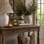 A rustic dark wood console table in a bright entryway, styled with a white ceramic table lamp, a green braided picture frame, stacked books, two large white vases with leafy plants, and a wicker basket holding a striped throw blanket.