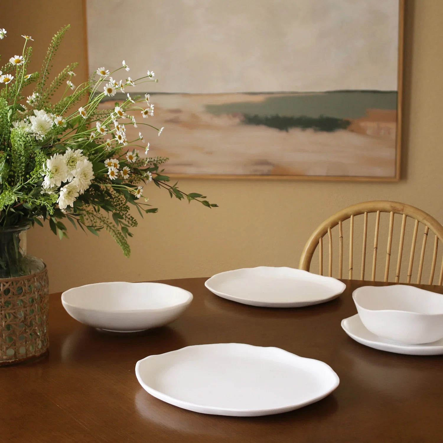 Dining table with white ceramic plates and bowls, vase of flowers, and a painting on the wall.