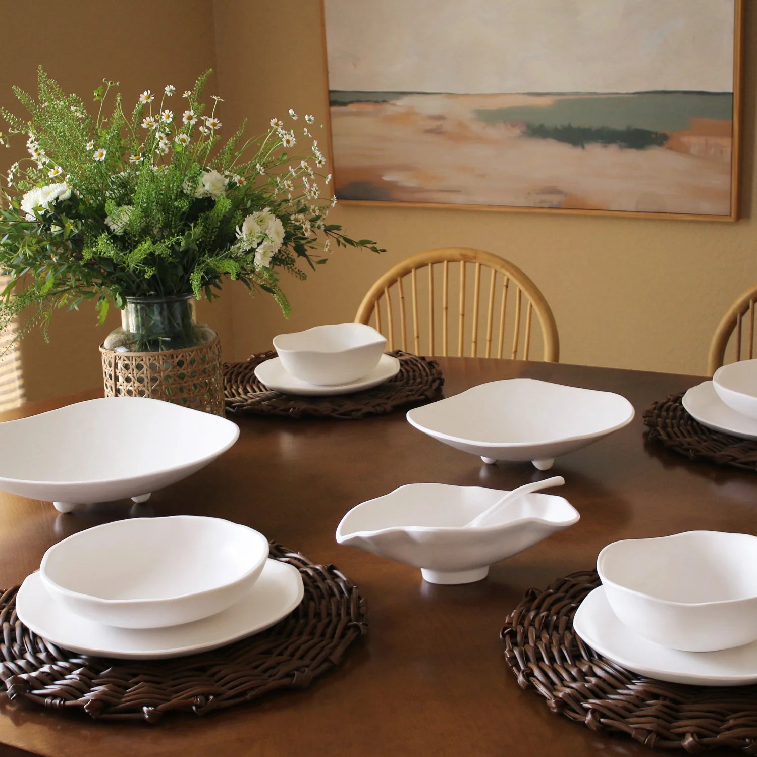Dining table set with white ceramic dishes on brown placemats, floral arrangement, and abstract painting in the background.