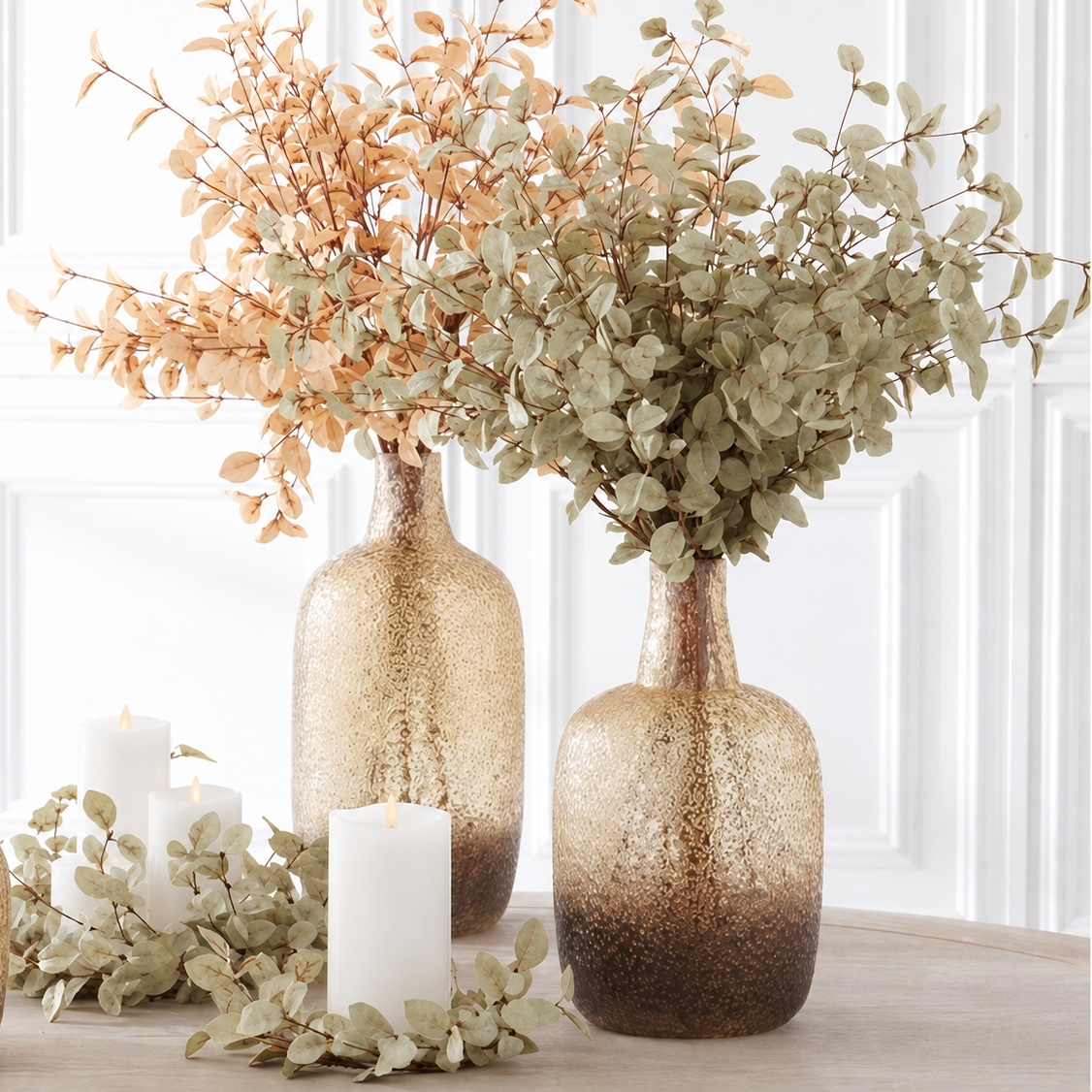 Three textured glass vase centers in a warm champagne-to-brown ombre finish, styled with dried botanicals and white candles on a light wood surface against a white paneled wall.