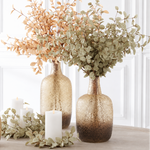 Three textured glass vase centers in a warm champagne-to-brown ombre finish, styled with dried botanicals and white candles on a light wood surface against a white paneled wall.