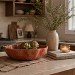 A rustic terracotta-colored ceramic bowl filled with artichokes sits on a beige woven placemat on a kitchen island. A tall textured vase with greenery, a lit candle in a glass holder, and stacked books are arranged nearby in a warm, modern kitchen setting.