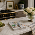 Kitchen counter with a book, bowl of artichokes, vase of white flowers, and decorative items.