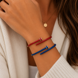 A close-up of a person's wrist wearing two slim bangle bracelets, one red and one blue, with small gold-toned connector details. The person is wearing a cream knit cardigan and a delicate gold pendant necklace.