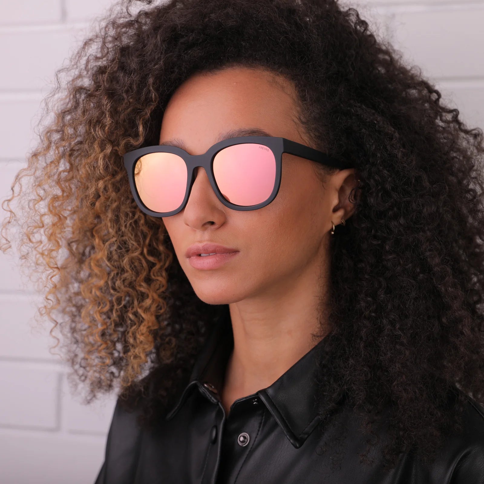 Woman wearing black sunglasses with pink lenses against a white brick wall.