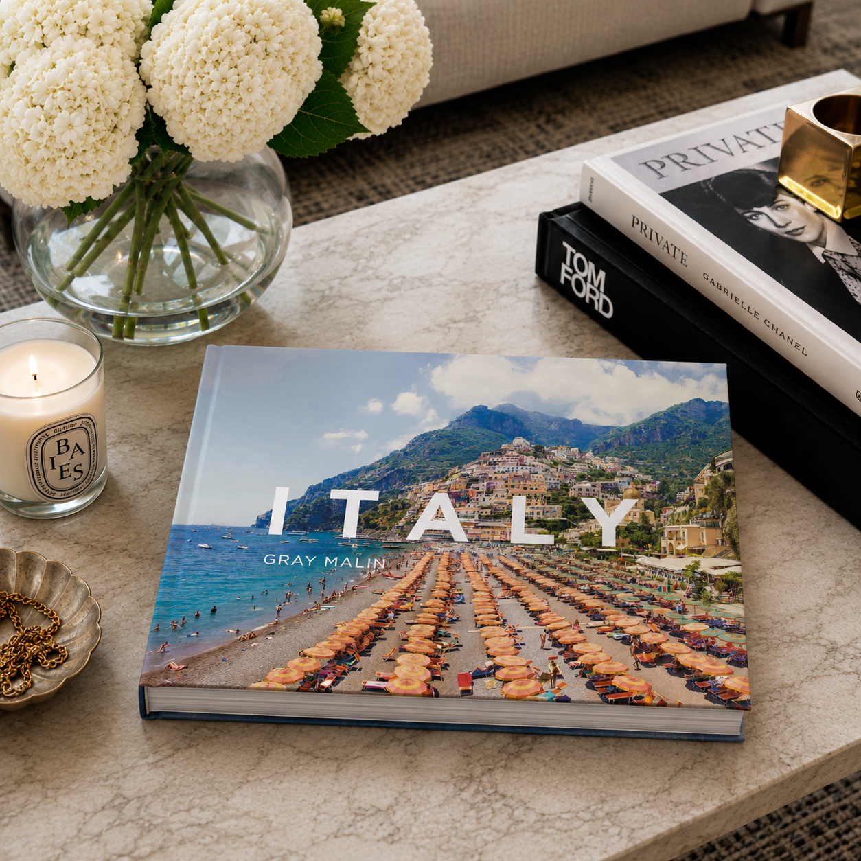 Book titled 'Italy' by Gray Malin on a coffee table with a candle and flowers.