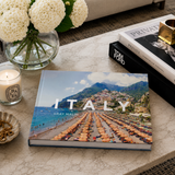 Book titled 'Italy' by Gray Malin on a coffee table with a candle and flowers.