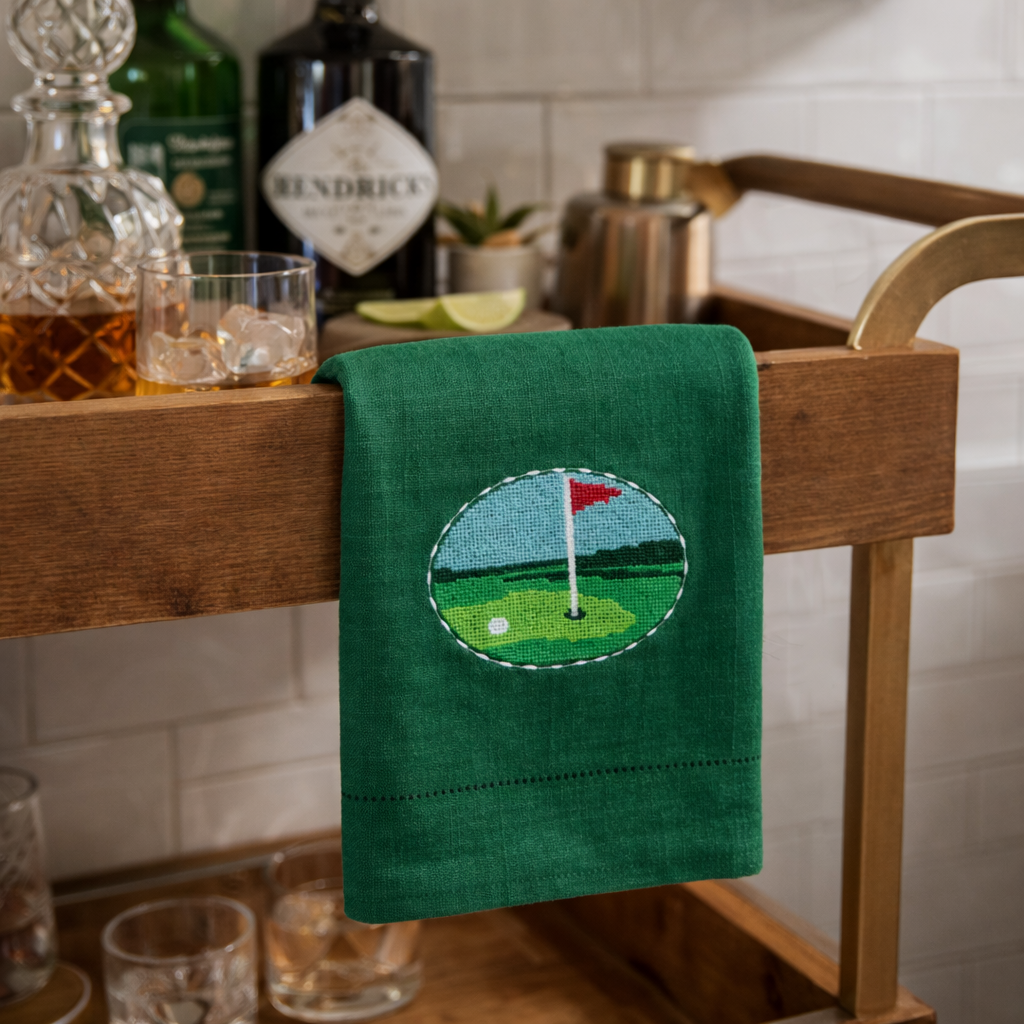Green towel with golf course design hanging on a wooden bar cart with drinks in the background.