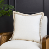 A square off-white throw pillow with a textured beige fringed border, resting on a white upholstered armchair with a dark wood cane armrest, set against a dark paneled wall with a green plant leaf in the corner.
