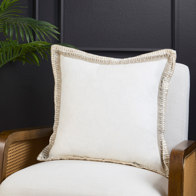 A square off-white throw pillow with a textured beige fringed border, resting on a white upholstered armchair with a dark wood cane armrest, set against a dark paneled wall with a green plant leaf in the corner.