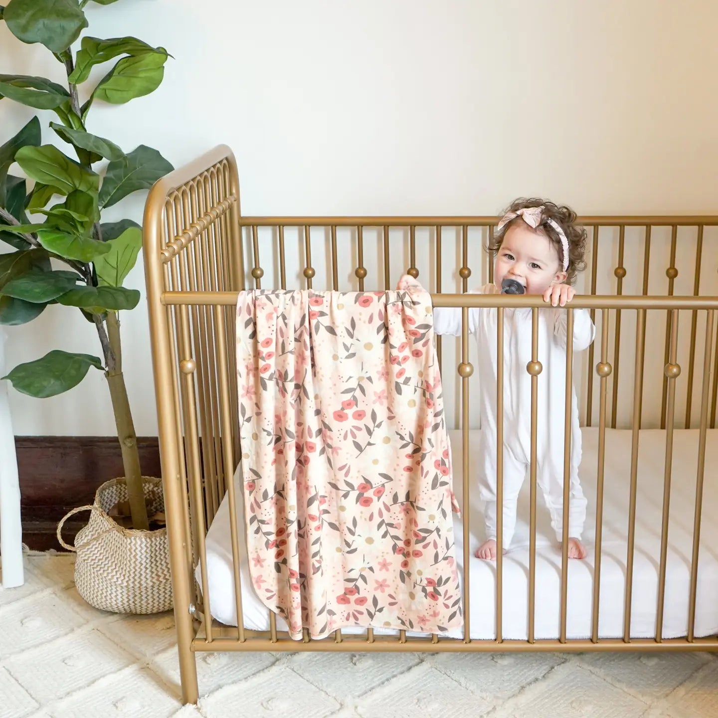 Child in a gold crib with a floral blanket, next to a plant