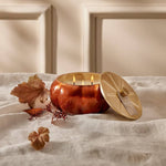 Copper pumpkin-shaped candle with a beige lid on a white surface with autumn leaves.