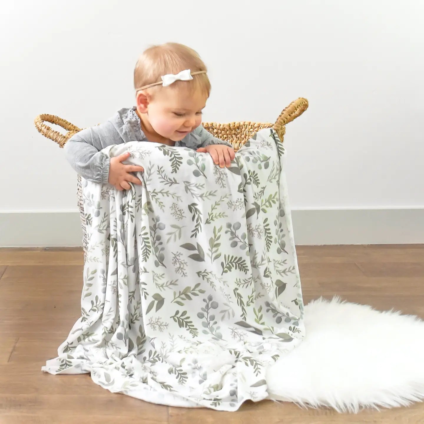 Baby holding a floral-patterned blanket in a wicker basket on a wooden floor.