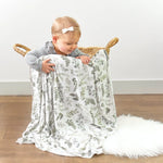 Baby holding a floral-patterned blanket in a wicker basket on a wooden floor.