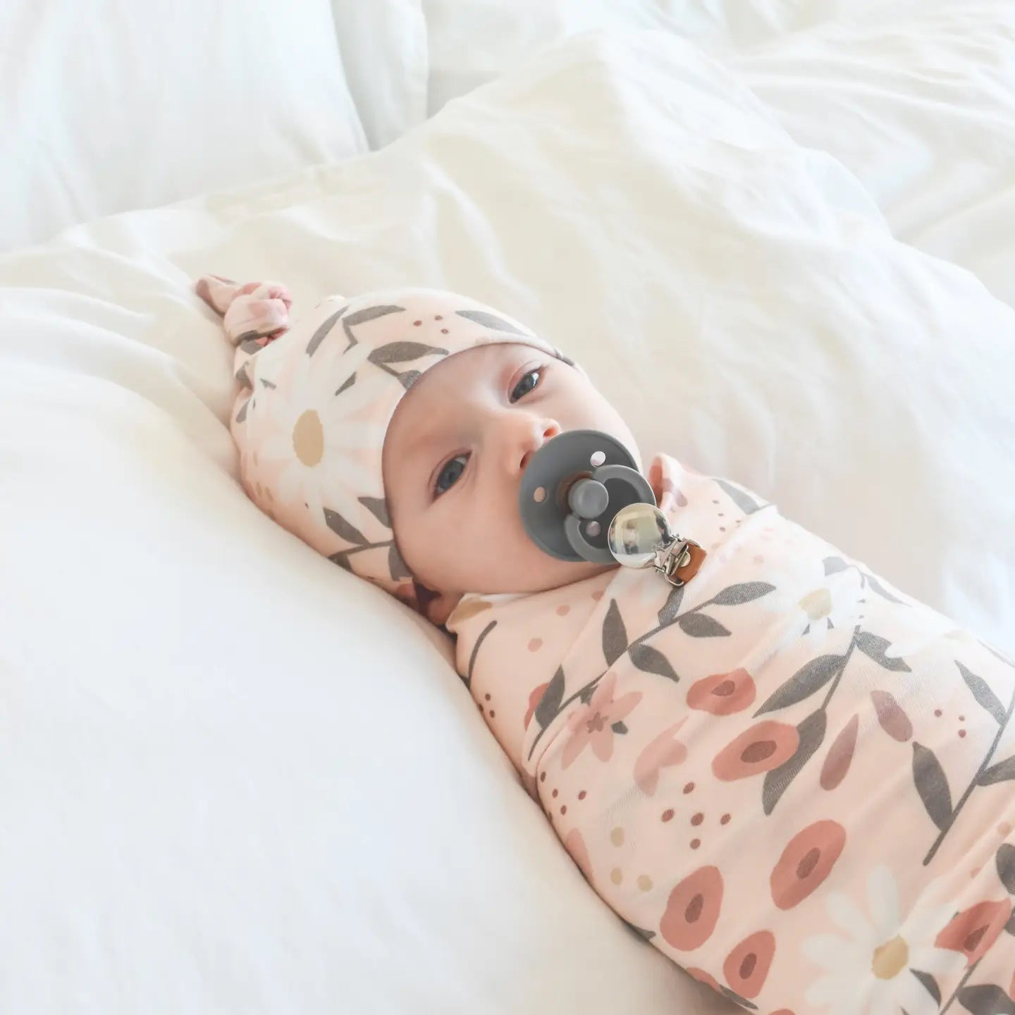 Baby swaddled in a floral blanket with a pacifier, lying on a white surface.