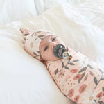 Baby swaddled in a floral blanket with a pacifier, lying on a white surface.