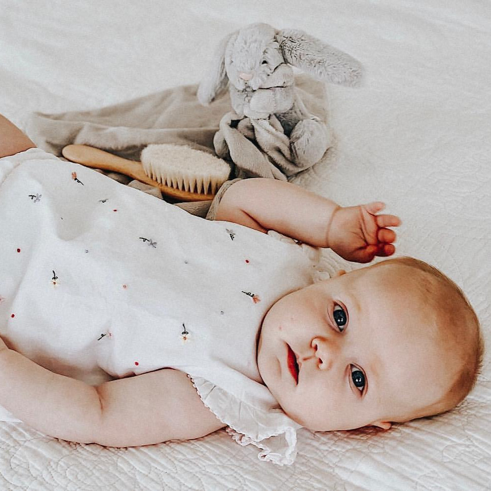 Baby lying on a white blanket with a toy and hairbrush nearby