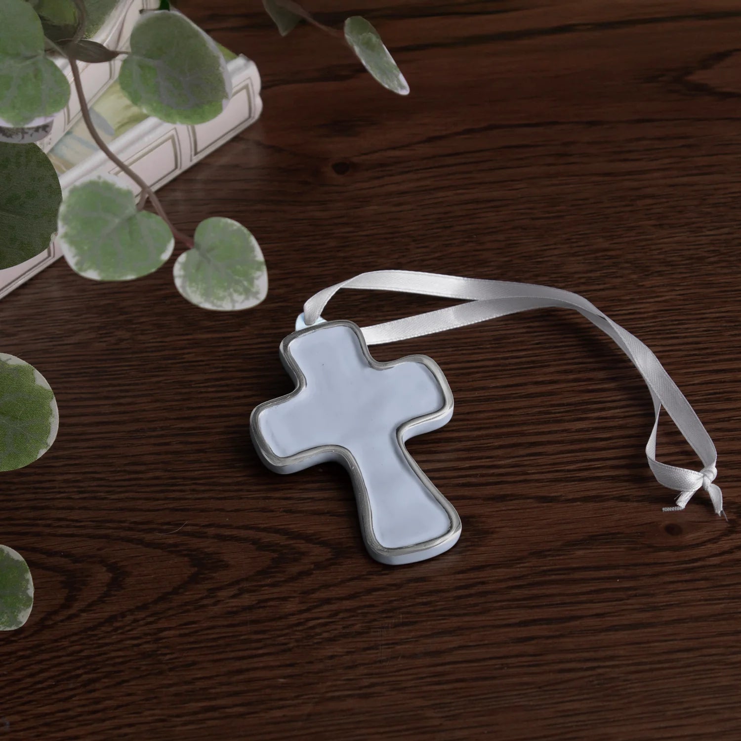 encanto athens cross ornament periwinkle with a white ribbon on a wooden surface with green leaves.