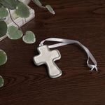 encanto athens cross ornament cream with a white ribbon on a wooden surface with green leaves.