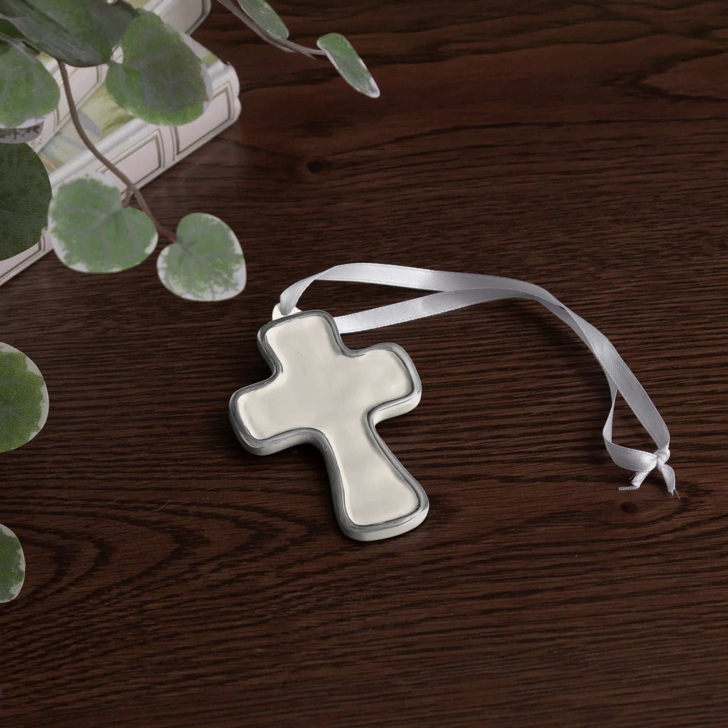 encanto athens cross ornament cream with a white ribbon on a wooden surface with green leaves.