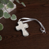 encanto athens cross ornament cream with a white ribbon on a wooden surface with green leaves.