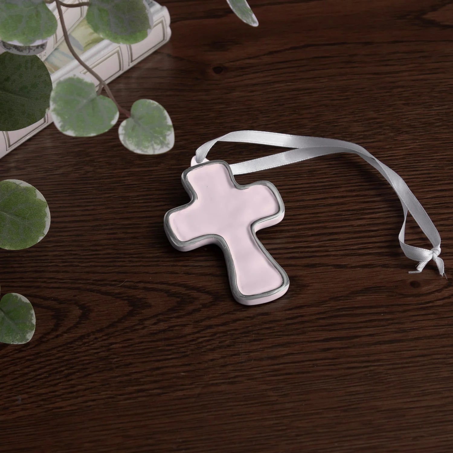 encanto athens cross ornament pink/camellia with a white ribbon on a wooden surface with green leaves.