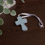 encanto athens cross ornament slate/blue with a white ribbon on a wooden surface with green leaves.