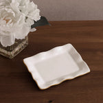 White ceramic dish with gold rim on a wooden surface next to a vase of white flowers.