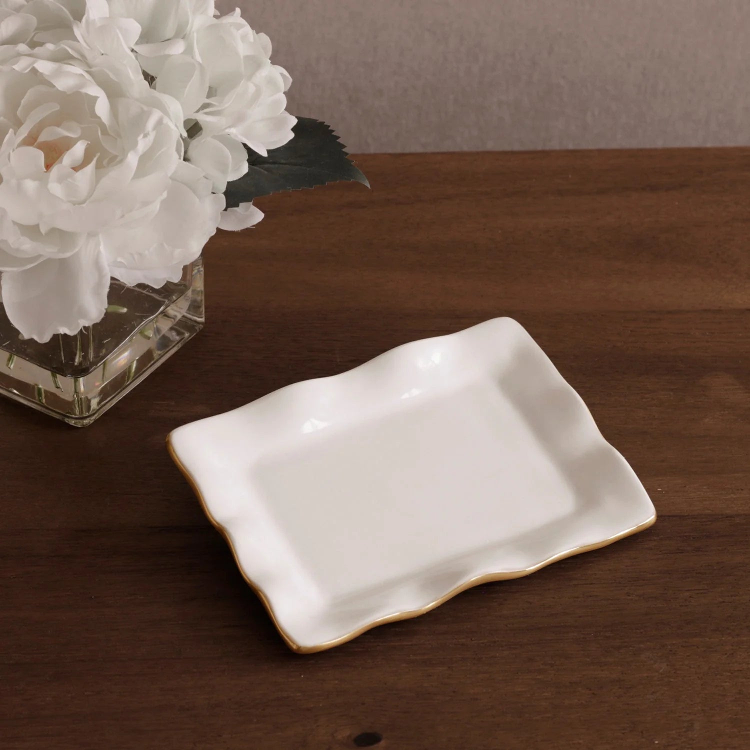 White ceramic dish with gold rim on a wooden surface next to a vase of white flowers.