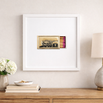 Framed artwork of a matchbox on a wall above a wooden shelf with decor items.