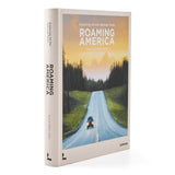 A hardcover book titled 'Roaming America: Exploring All the National Parks' standing upright on a white background. The cover features a scenic photograph of a winding road through a forest under a warm sunset sky, with the title in bold white text.