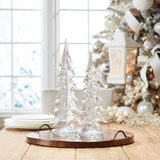 Decorative glass Christmas trees on a wooden table with a Christmas tree in the background.