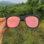 Sunglasses with pink lenses held in front of a green, natural background