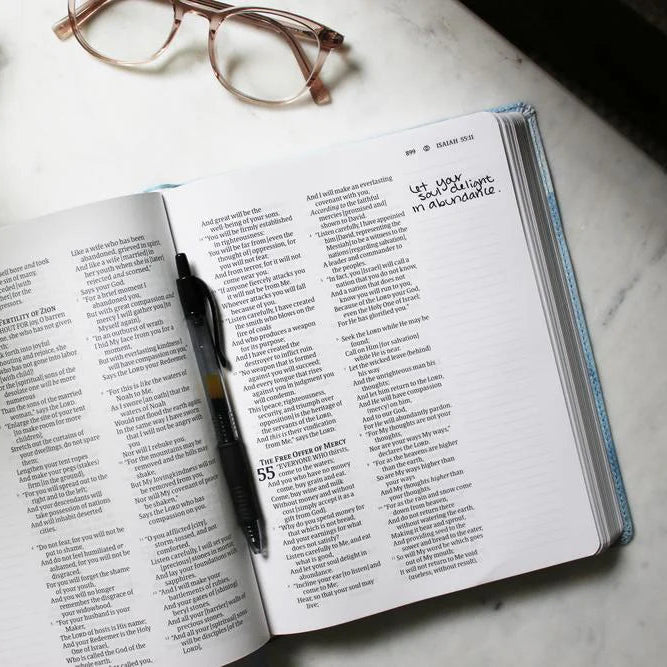 Open Anne Neilson Bible with a pen on a marble surface, next to glasses and a plant.