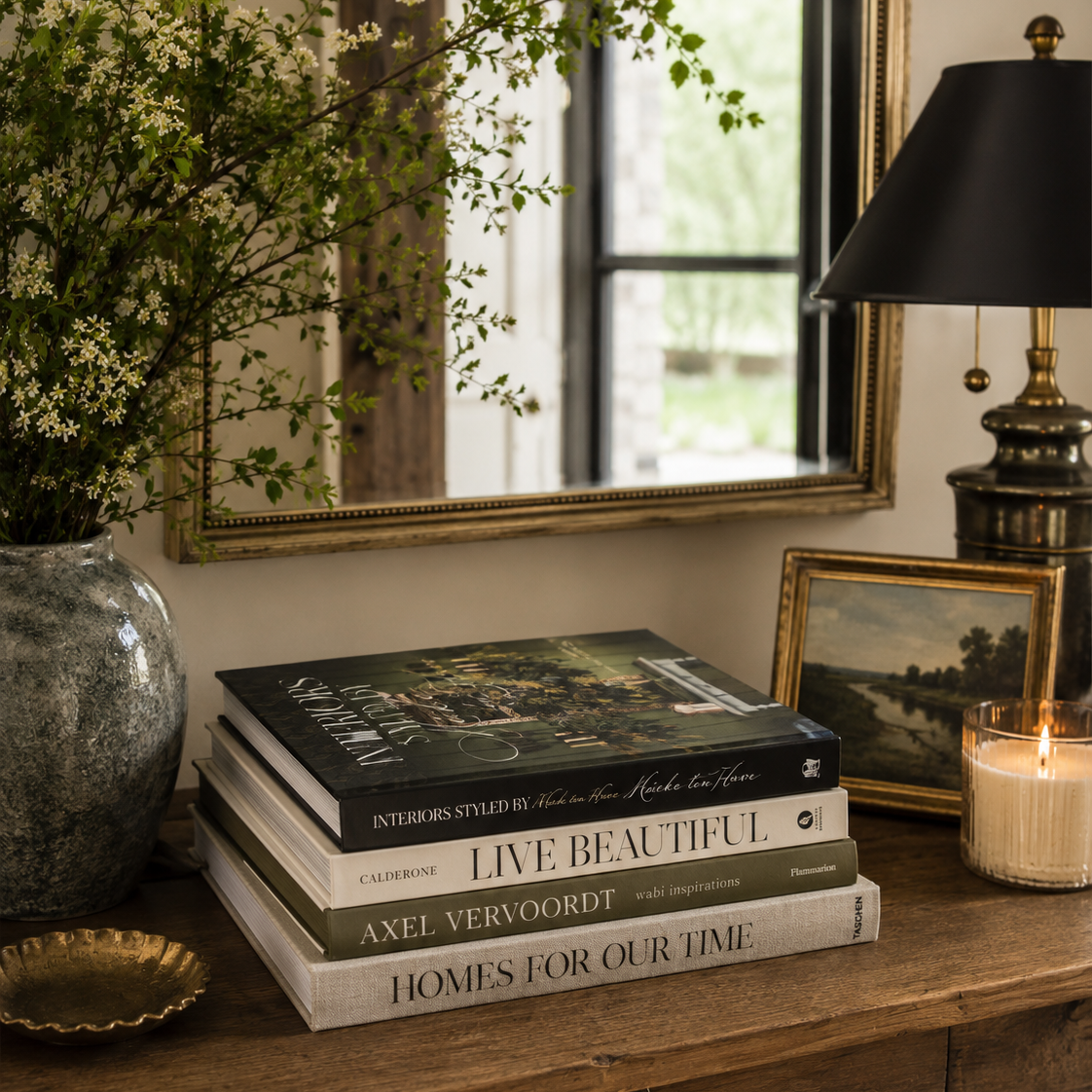 Stack of books on a wooden table with a vase, lamp, and mirror in the background