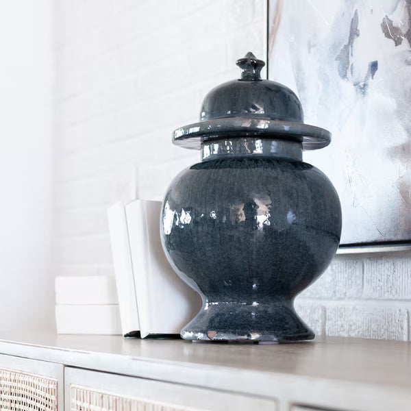 Decorative dark grey urn with a lid on a shelf against a white wall.