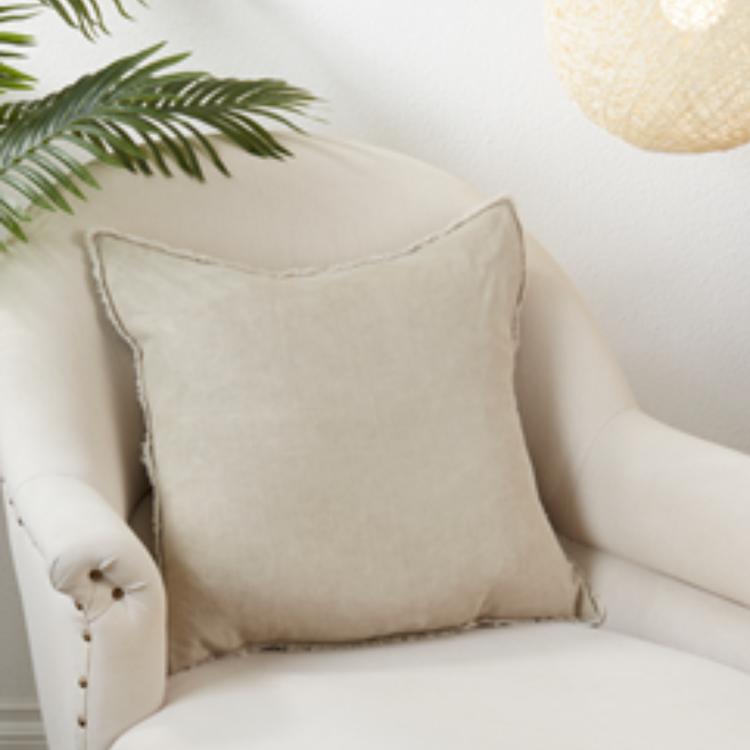 A square beige throw pillow with frayed edges resting on a cream-colored armchair, with a green plant leaf and a woven pendant light visible in the background.