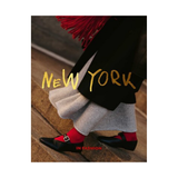 A fashion editorial-style photo showing the lower half of a person wearing a black coat, light gray skirt, bright red socks, and black Mary Jane shoes on stone steps. Large gold text reads “NEW YORK,” with smaller red text near the bottom reading “IN FASHION". hardcover coffee table book on white background
