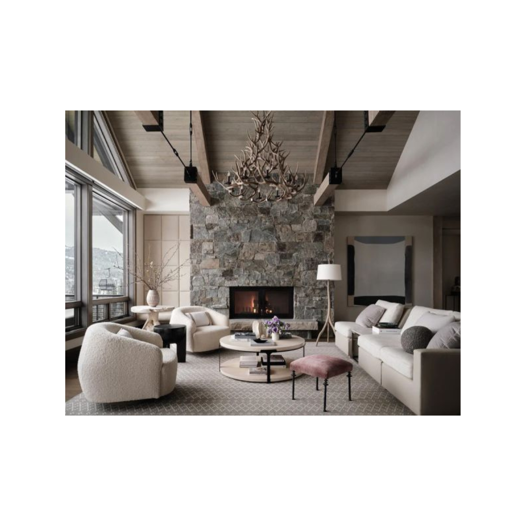 A luxury modern living room featuring a large stone fireplace, a high wood-beamed ceiling, a dark metal chandelier, a textured wool armchair, a cream sofa, and a pink upholstered ottoman from hardcover book "world's best: 50 interiors from around the globe" on a white background