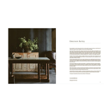 An open book view of interior design spread with a dark, moody room scene on the left showing a rustic wooden sideboard and table with plants, and a text column on the right headed Emerson Bailey.