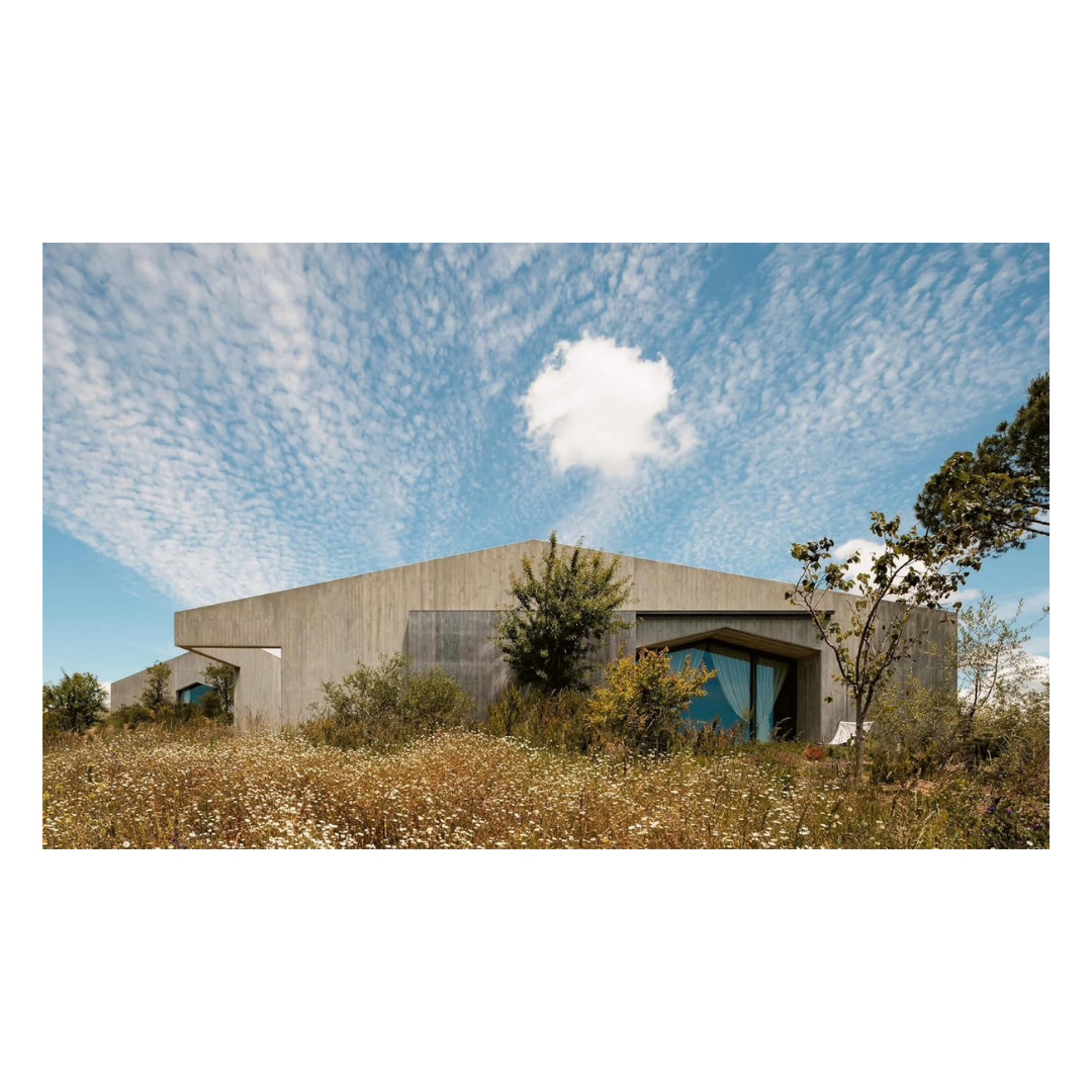 A rectangular art print showing a modern concrete building nestled in dry grass under a bright blue sky with wispy white clouds from the hardcover book "contemporary living yearkbook 2026" on a white background